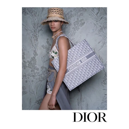 A tanned woman in a summer-style ensemble holds a branded tote bag against a floral backdrop, evoking high-fashion and vacation chic.
