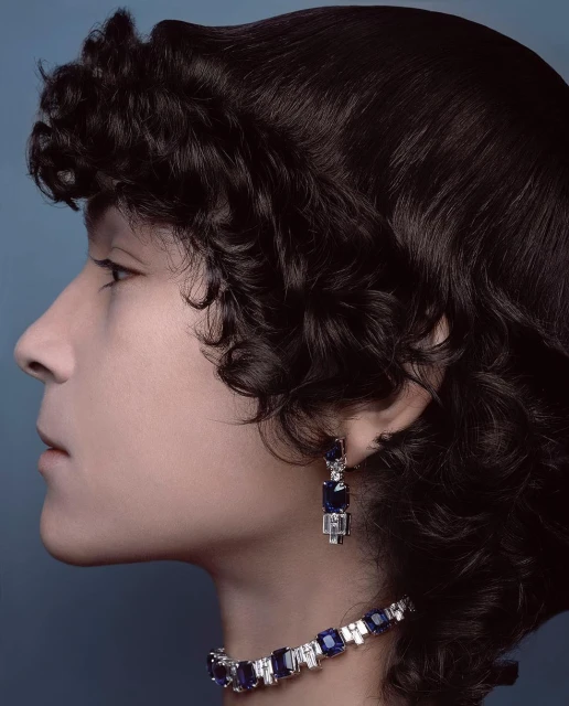 Profile of a woman with dark curly hair wearing diamond and sapphire jewelry against a pale blue background in a beauty shot.