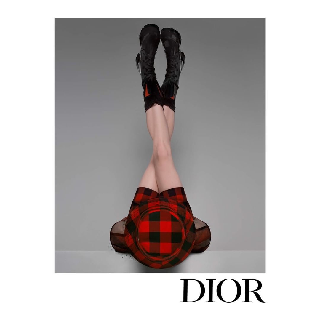 Fashion shot of a woman's lower body wearing black boots, plaid skirt and hat, with legs raised against a gray background. Designer brand Dior.