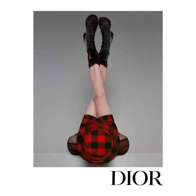 Fashion shot of a woman's lower body wearing black boots, plaid skirt and hat, with legs raised against a gray background. Designer brand Dior.