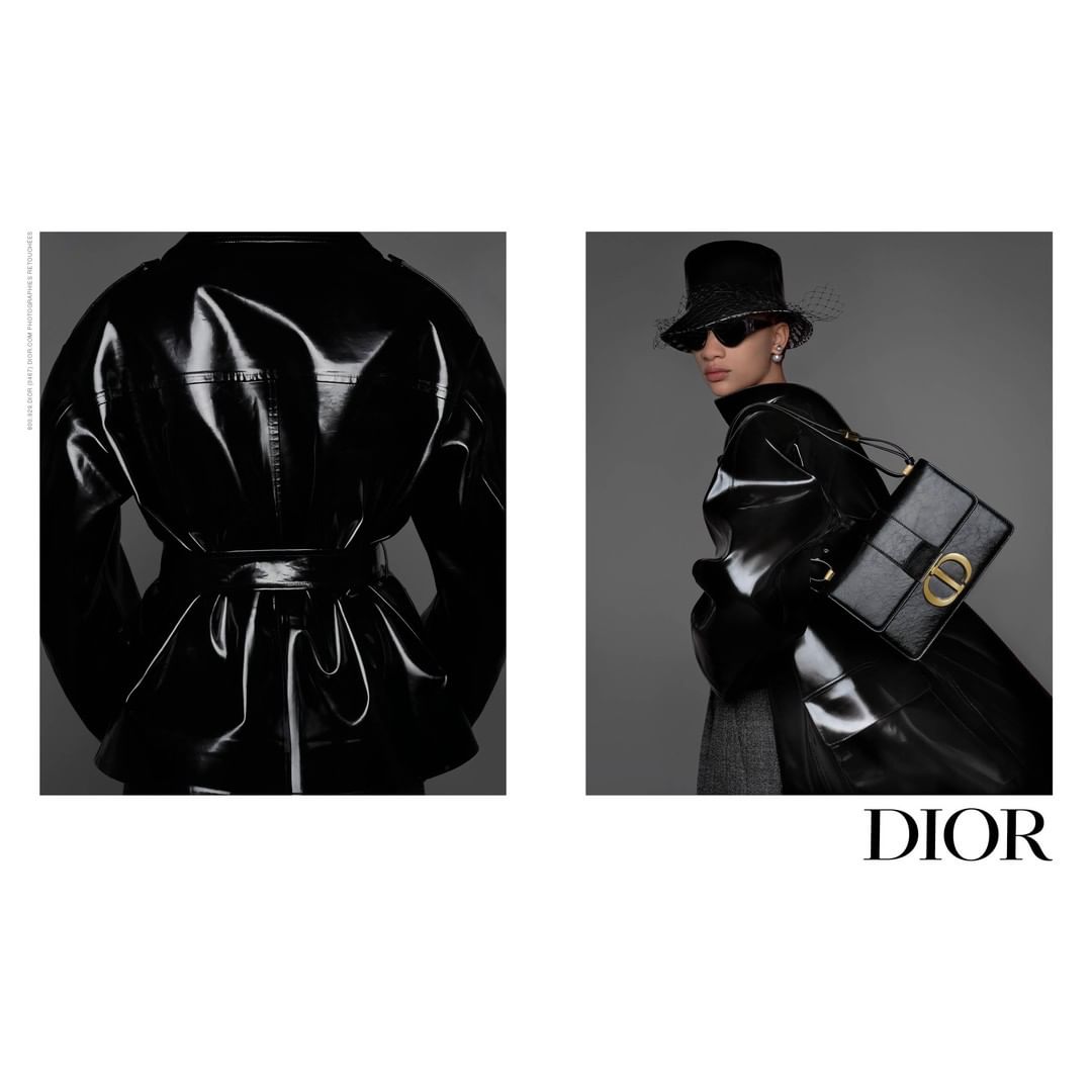 Fashion shot of a woman wearing a glossy black trench coat, bucket hat, and sunglasses, carrying a black Dior purse against a gray background.
