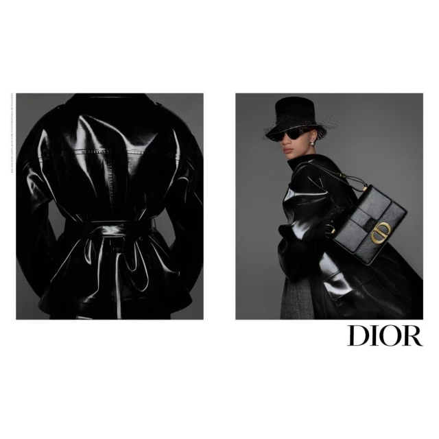 Fashion shot of a woman wearing a glossy black trench coat, bucket hat, and sunglasses, carrying a black Dior purse against a gray background.