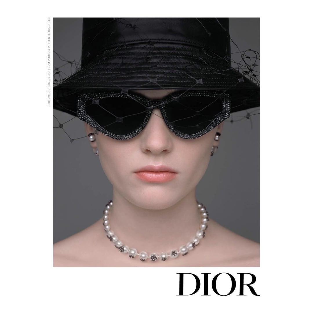 Headshot of a woman in a black hat and sunglasses, wearing a pearl necklace and earrings, posing in front of a gray backdrop. 
