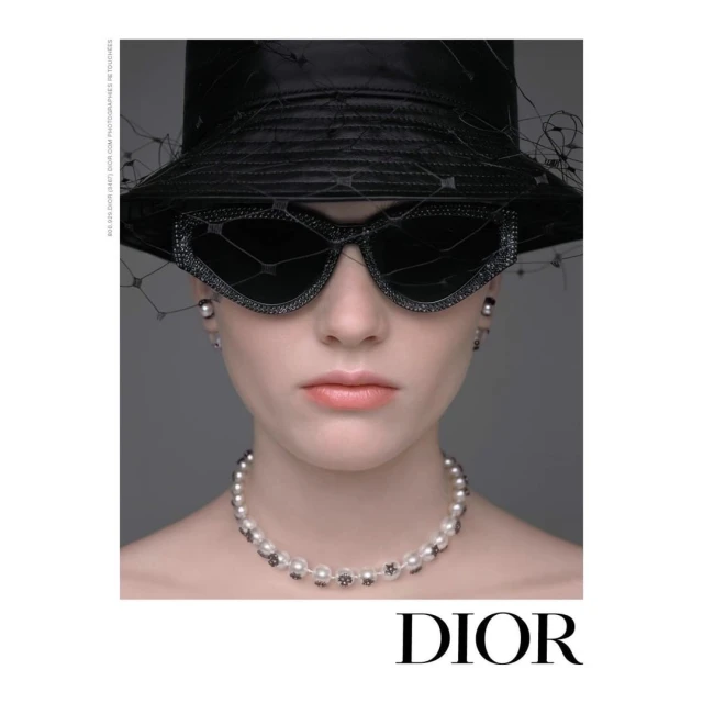 Headshot of a woman in a black hat and sunglasses, wearing a pearl necklace and earrings, posing in front of a gray backdrop. 