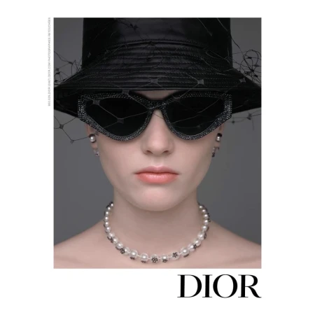 Headshot of a woman in a black hat and sunglasses, wearing a pearl necklace and earrings, posing in front of a gray backdrop. 