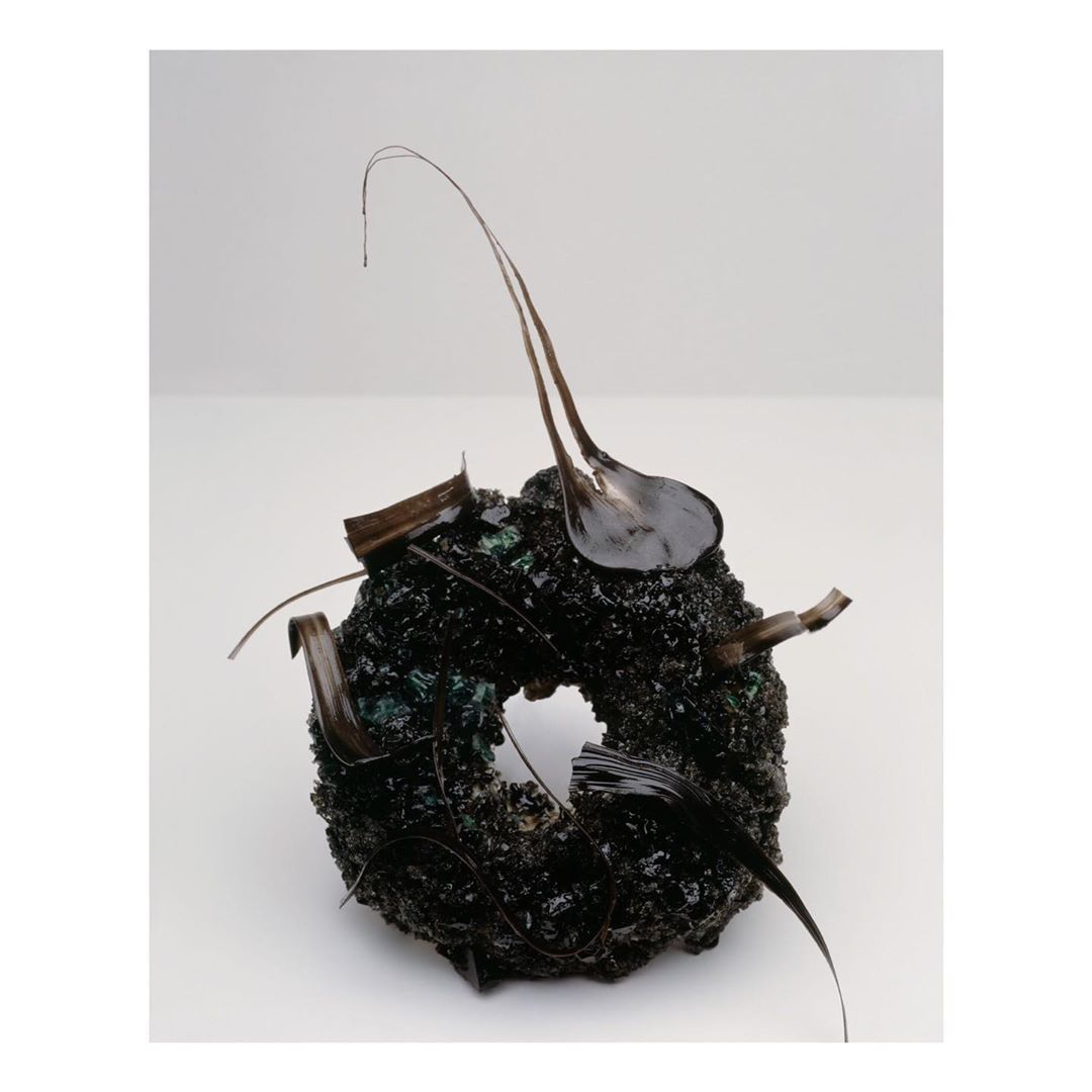 Abstract art piece featuring a dark, textured, donut-shaped object adorned with glossy, dark-colored, organic elements, against a plain background.