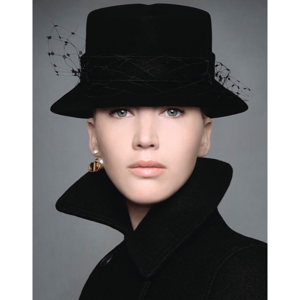 Portrait of a woman wearing a black structured hat with veil and a high-collared black coat against a neutral grey background