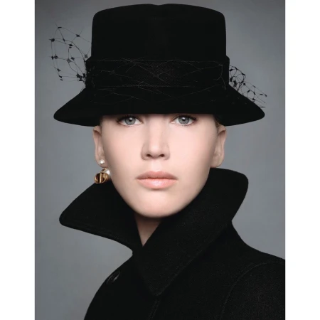 Portrait of a woman wearing a black structured hat with veil and a high-collared black coat against a neutral grey background