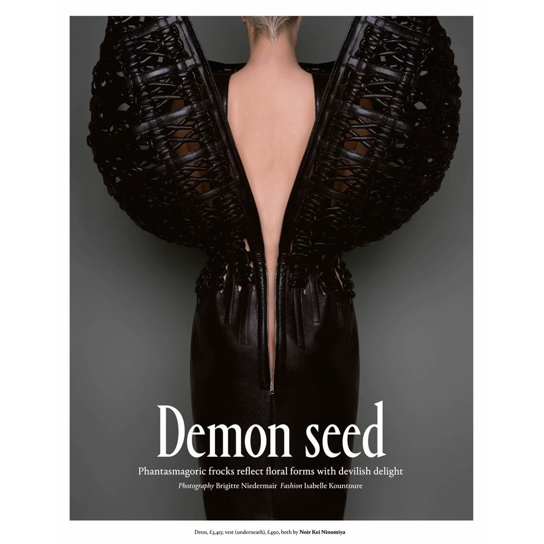 A woman models a black avant-garde dress with architectural wing-like shoulders against a grey background in a fashion editorial shot.