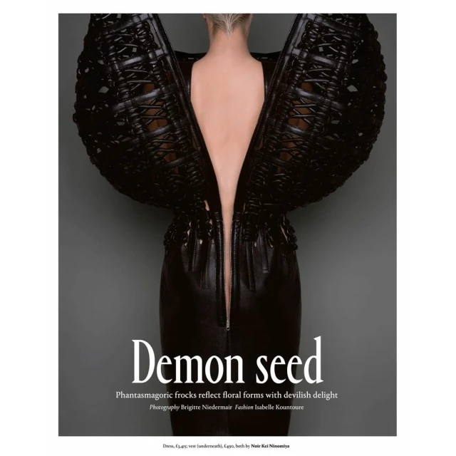 A woman models a black avant-garde dress with architectural wing-like shoulders against a grey background in a fashion editorial shot.