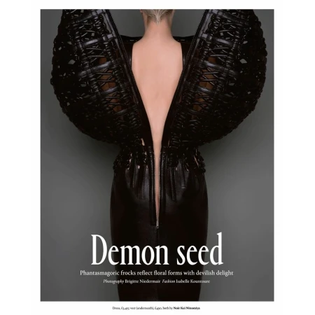 A woman models a black avant-garde dress with architectural wing-like shoulders against a grey background in a fashion editorial shot.