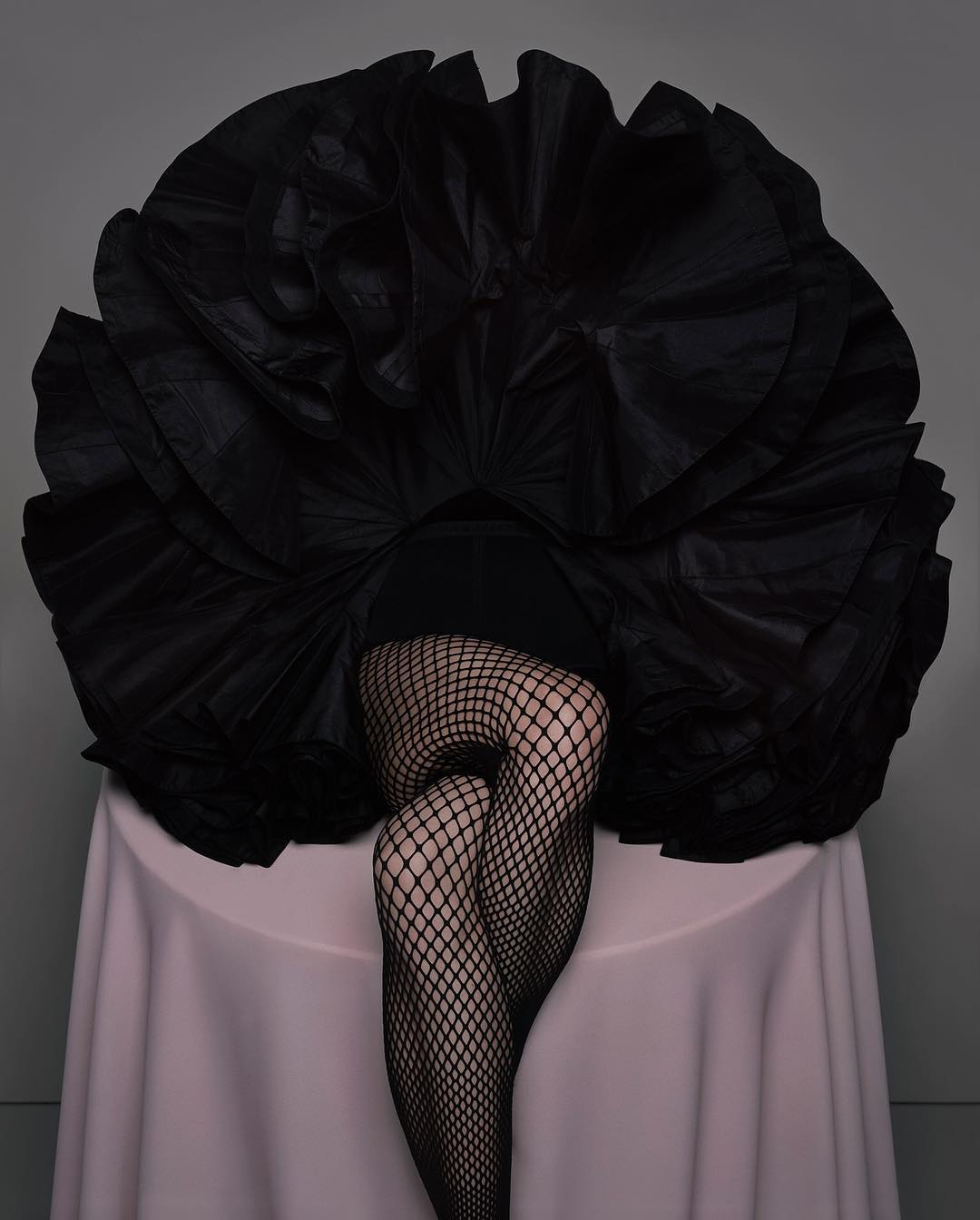 A person's legs in fishnet stockings are visible under a black ruffled dress atop a pink table, set against a gray backdrop.
