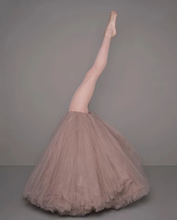Surreal portrait of a dancer wearing a dusty rose tutu with one leg extended straight up into the air against a neutral grey background
