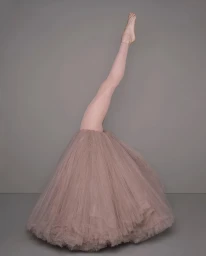 Surreal portrait of a dancer wearing a dusty rose tutu with one leg extended straight up into the air against a neutral grey background