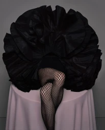 Legs in fishnet tights extending beneath a massive, sculptural black ruffled garment draped over a table