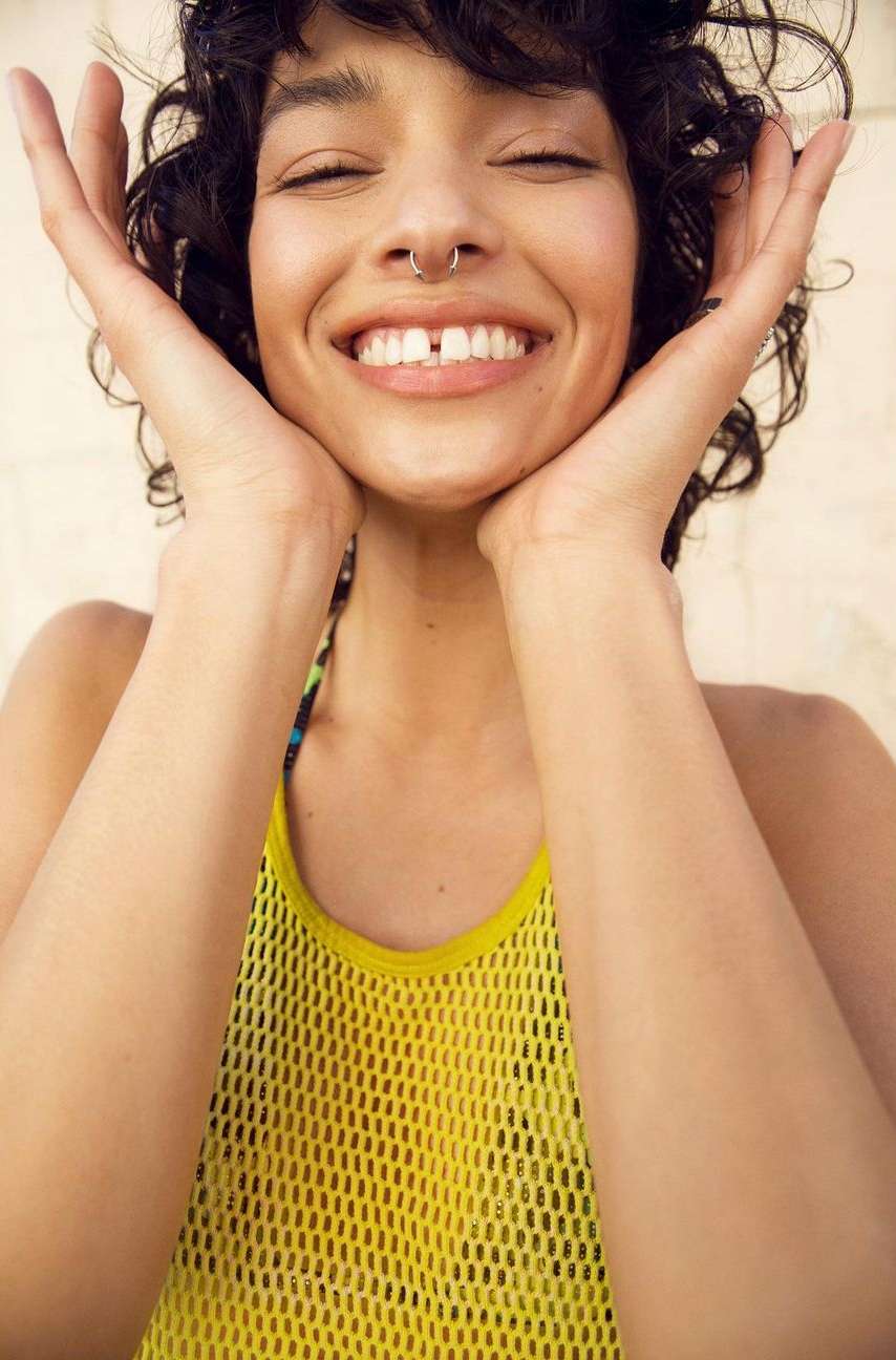 Portrait of a smiling woman with dark curly hair and a yellow mesh top, eyes closed, and hands near her face, in a bright indoor setting.