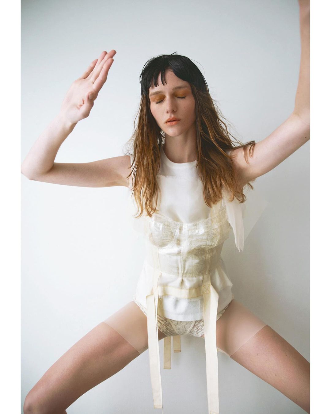 A single adult female in a white top and panty stands in a light studio, her arms are raised, posing with a neutral facial expression.