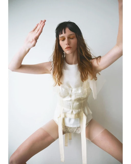 A single adult female in a white top and panty stands in a light studio, her arms are raised, posing with a neutral facial expression.