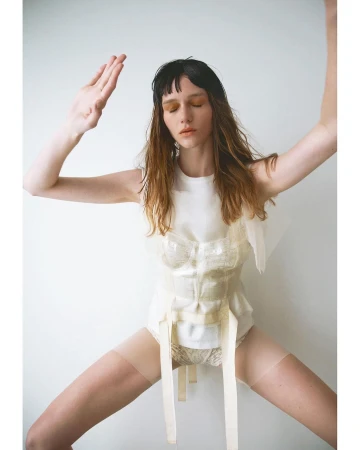 A single adult female in a white top and panty stands in a light studio, her arms are raised, posing with a neutral facial expression.