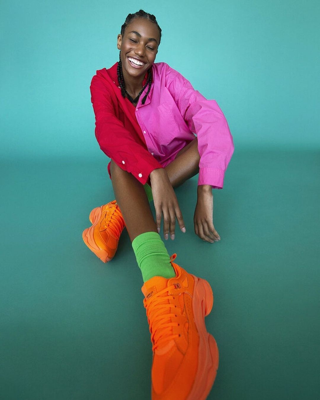 A young woman with braids smiles sitting on a green background wearing bright orange sneakers, a half pink half red shirt, and green socks.