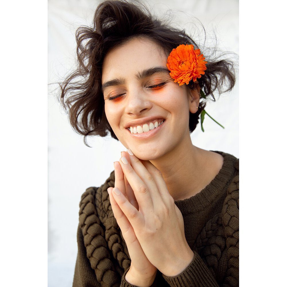 Portrait of a smiling woman with a flower in her hair, wearing a brown sweater against a white background in a studio setting.