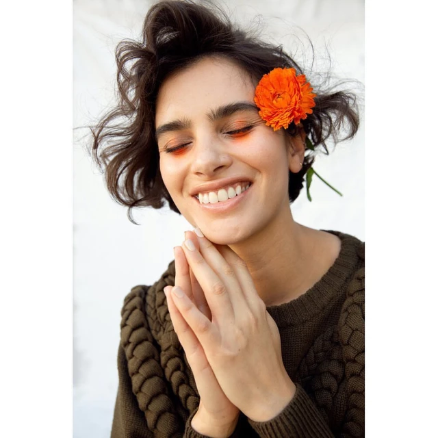 Portrait of a smiling woman with a flower in her hair, wearing a brown sweater against a white background in a studio setting.