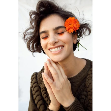 Close-up portrait of a smiling woman with dark short hair holding hands near her chin, wearing a textured olive sweater and an orange flower tucked behind her ear.