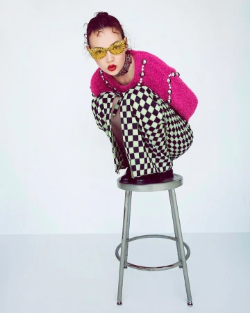 Model wearing hot pink sweater and checkered pants crouching on a metal stool against a white background