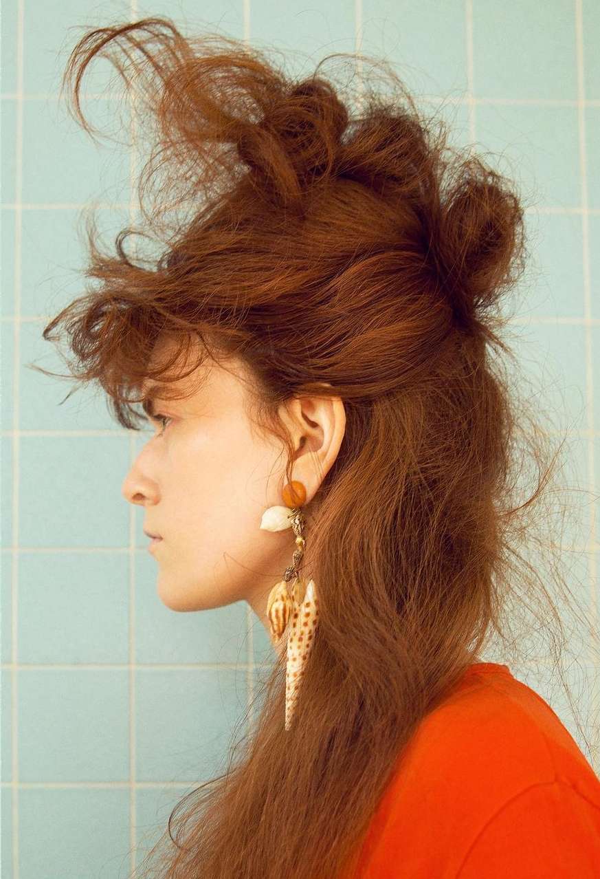 Profile shot of a woman with red hair wearing large seashell earrings against a blue tiled wall, showing a fashion and beauty aesthetic.