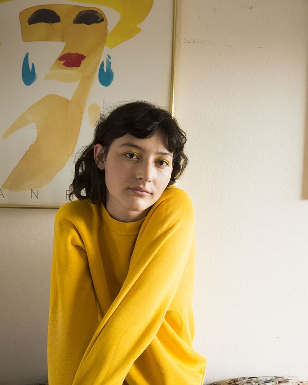A portrait of an adult female with short brown hair wearing a yellow sweater, posed in front of a vintage poster indoors.