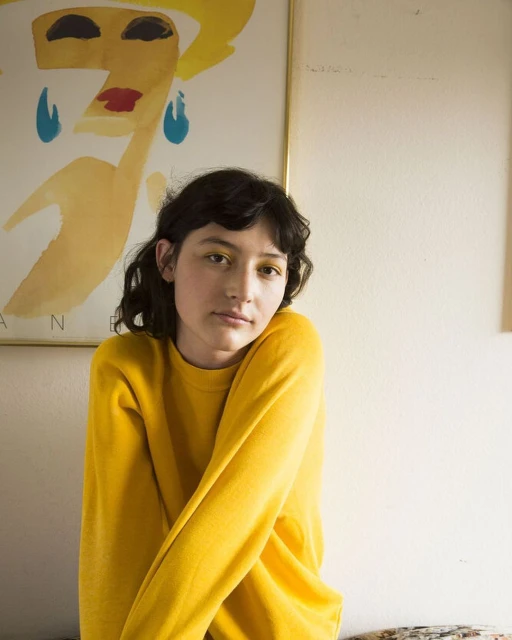 A portrait of an adult female with short brown hair wearing a yellow sweater, posed in front of a vintage poster indoors.