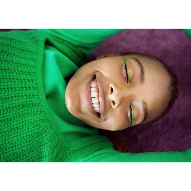 A smiling woman with bright green eyeliner, wearing a green sweater, is laying on a dark purple surface in a portrait orientation.
