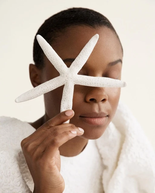Adult woman covers her eye with a white starfish in an indoor shot against a white background, wearing a white textured top.