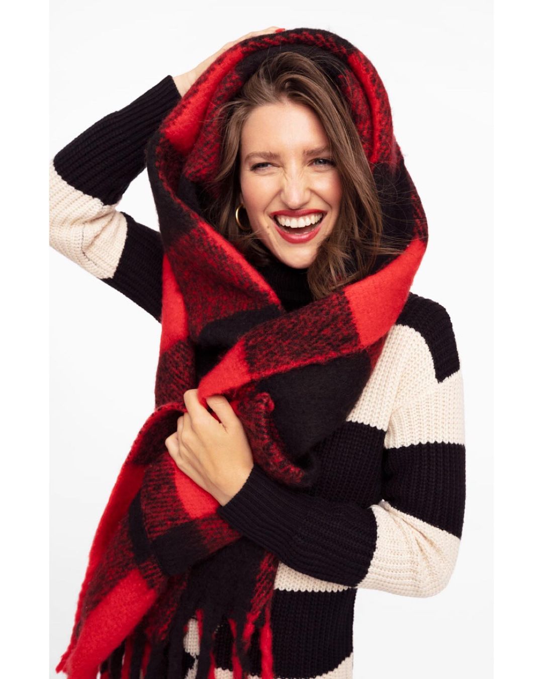 A woman smiles wearing a red and black plaid scarf as a hood, and a black and white striped sweater in a bright indoor setting.