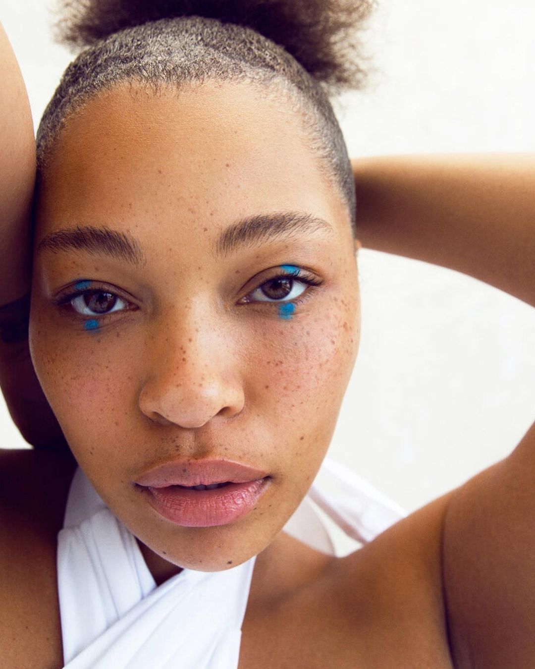 Close-up portrait of a young adult woman with brown hair, natural skin, and bright blue star-shaped makeup under her eyes against a white background.