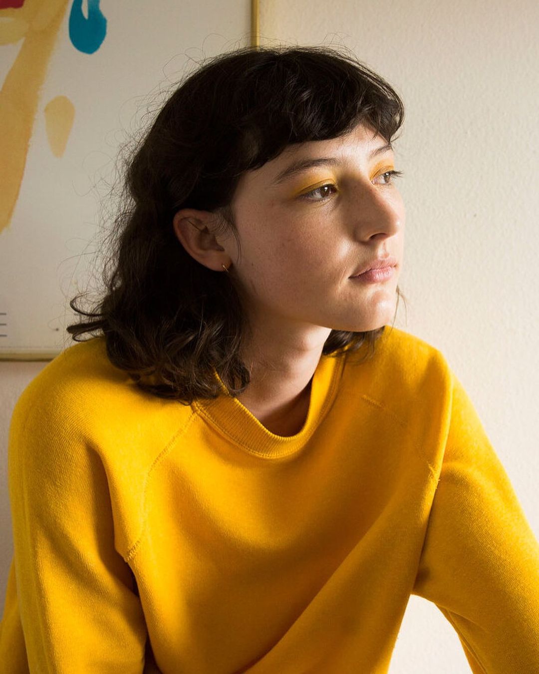 Portrait of a young adult woman with dark hair and yellow eyeshadow wearing a yellow sweater in an indoor setting with artwork.