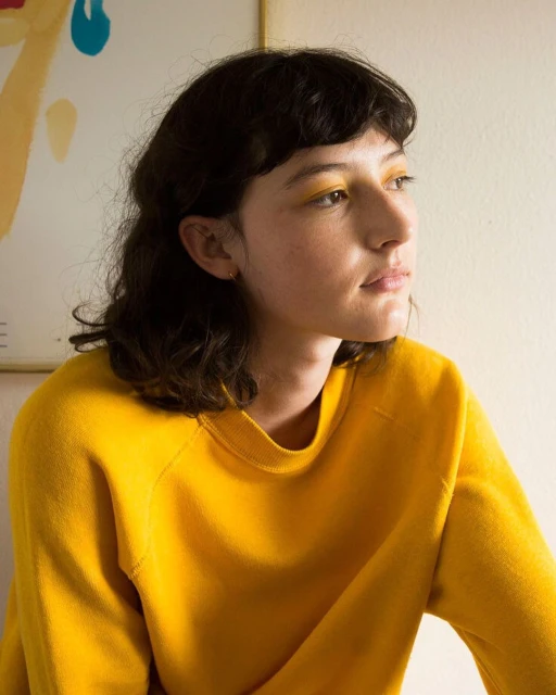 Portrait of a young adult woman with dark hair and yellow eyeshadow wearing a yellow sweater in an indoor setting with artwork.