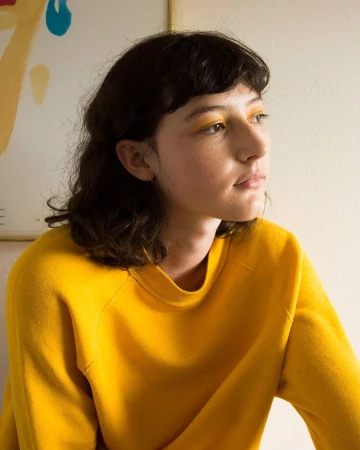 Portrait of a young adult woman with dark hair and yellow eyeshadow wearing a yellow sweater in an indoor setting with artwork.