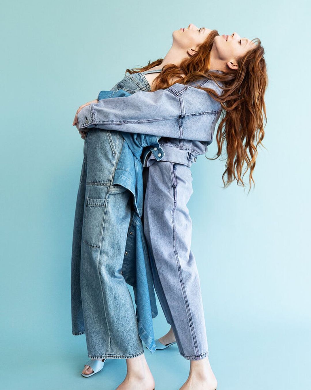 Two women in light blue denim outfits stand back-to-back in a fashion studio, looking upwards against a light blue backdrop.