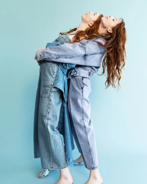 Two women in light blue denim outfits stand back-to-back in a fashion studio, looking upwards against a light blue backdrop.