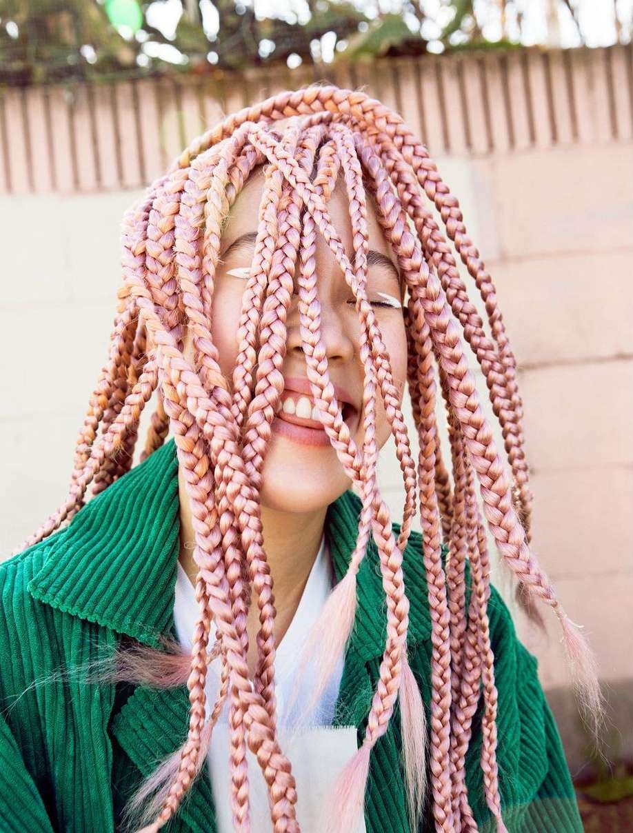 Portrait of a smiling woman with pink box braids wearing a green corduroy jacket, standing outside with a concrete wall background.