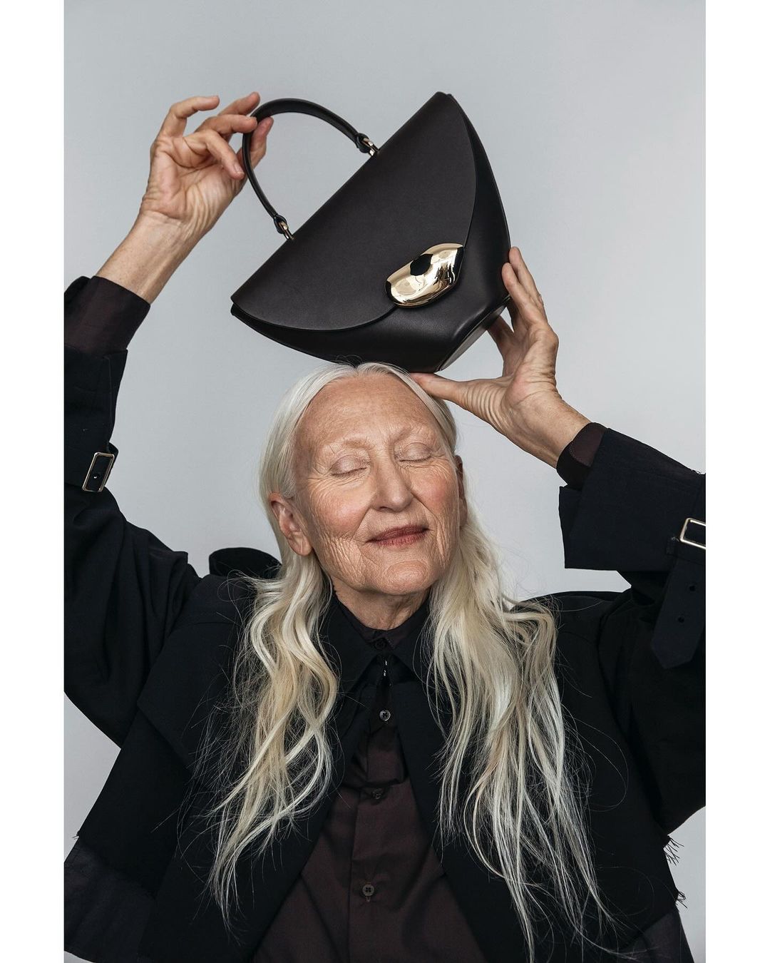 Elegant woman with closed eyes balances a black leather handbag with a gold clasp on her head in a studio shot.