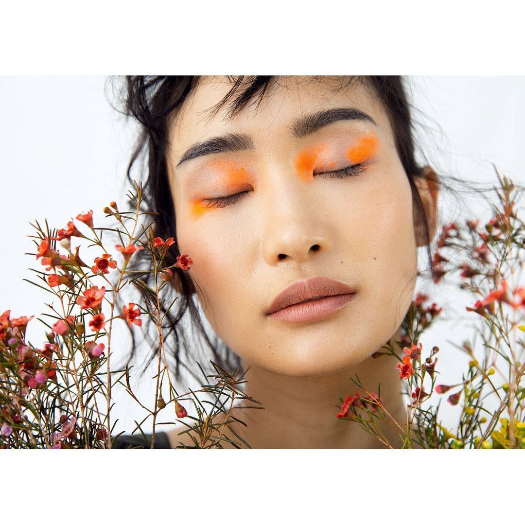 Close-up portrait of an Asian woman with closed eyes and vibrant orange eyeshadow, surrounded by delicate red and yellow wildflowers against white.