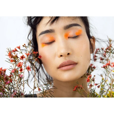 Close-up portrait of an Asian woman with closed eyes and vibrant orange eyeshadow, surrounded by delicate red and yellow wildflowers against white.