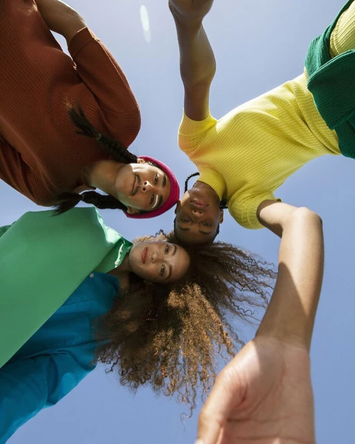 Low angle view of three young adults with colourful tops standing in a circle on a sunny day with clear blue sky above them.
