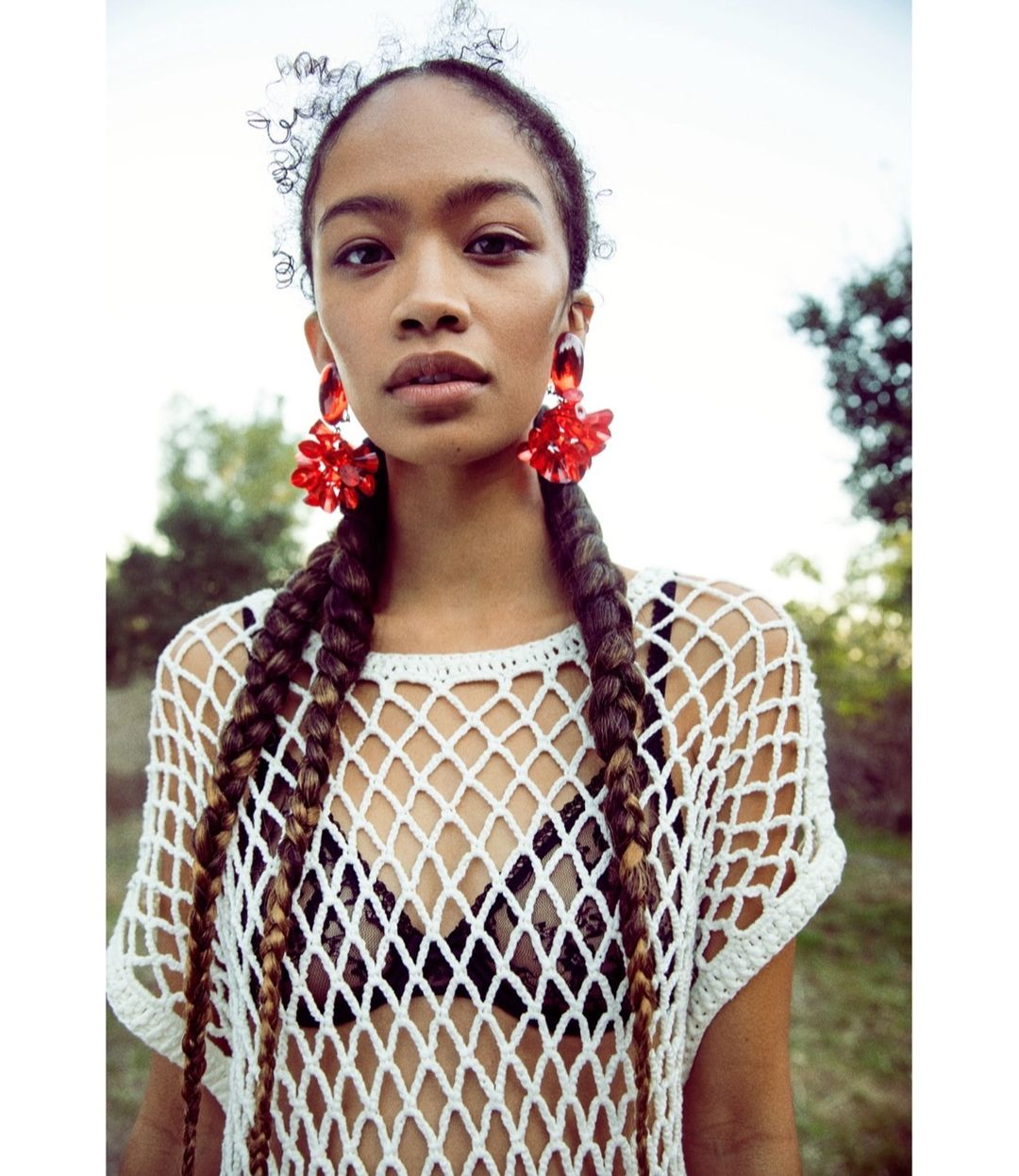 Woman with braided hair poses in a netted top with red earrings outdoors in natural light; fashion, beauty, and celebrity & entertainment.