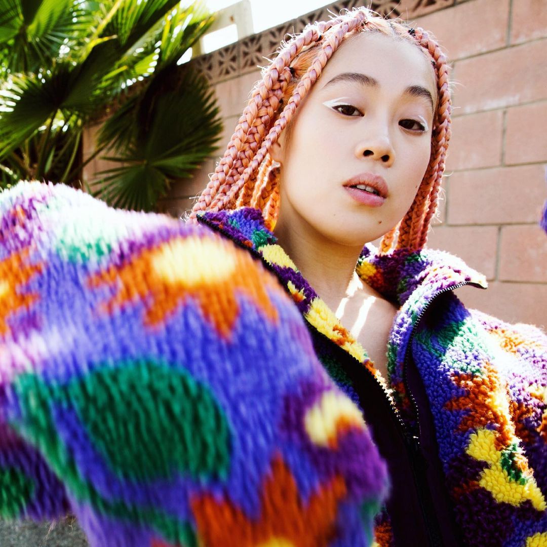 Portrait of a woman with peach-colored braids wearing a colorful patterned jacket. She is posing in front of a brick wall.