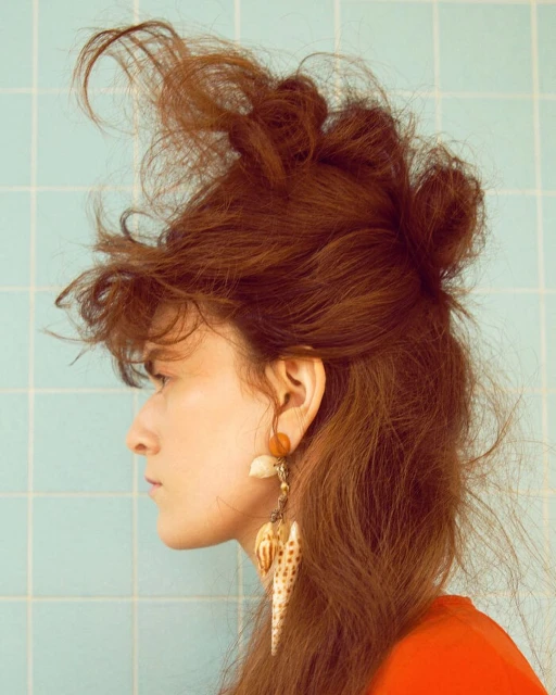 Profile portrait of a young adult female with auburn hair styled upwards, shell earrings, wearing an orange top, against a blue tile wall.