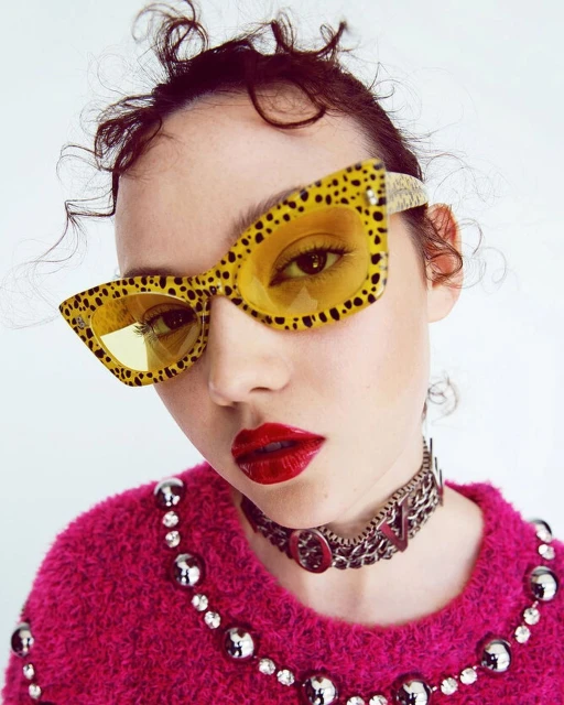 Close-up of a woman wearing yellow and black sunglasses with red lipstick and a studded choker against a white background.