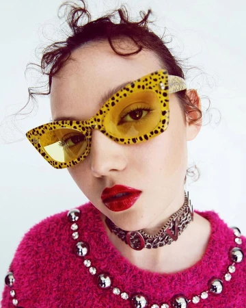 Close-up of a woman wearing yellow and black sunglasses with red lipstick and a studded choker against a white background.